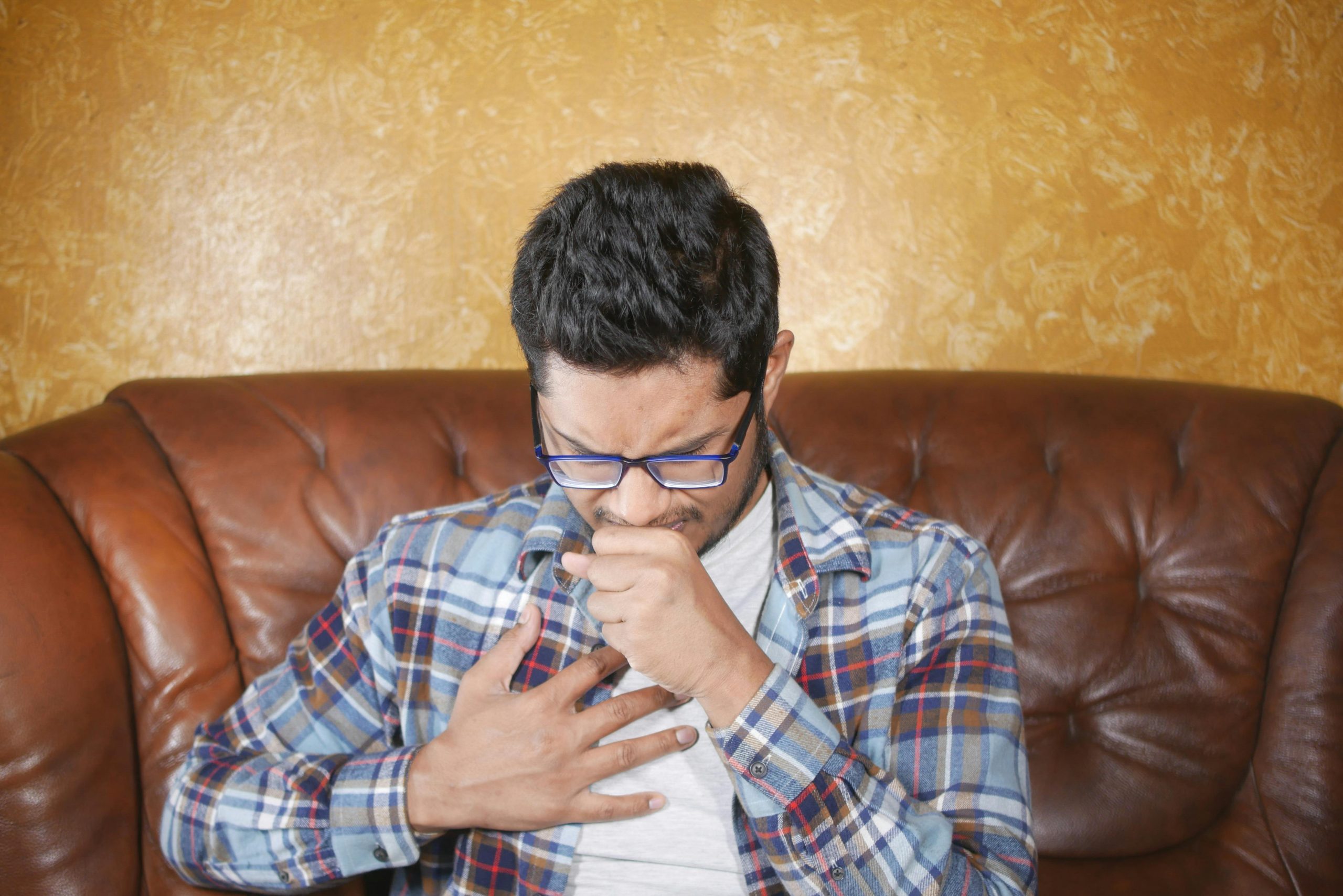 man sitting on a couch, coughing into one hand while the other hand rests on his chest