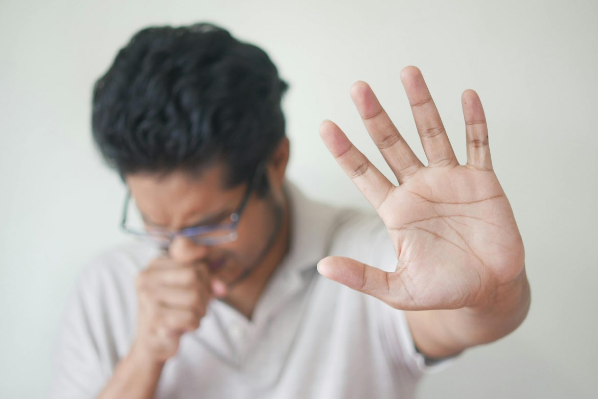 man coughing into one hand while raising the other hand in a stop gesture